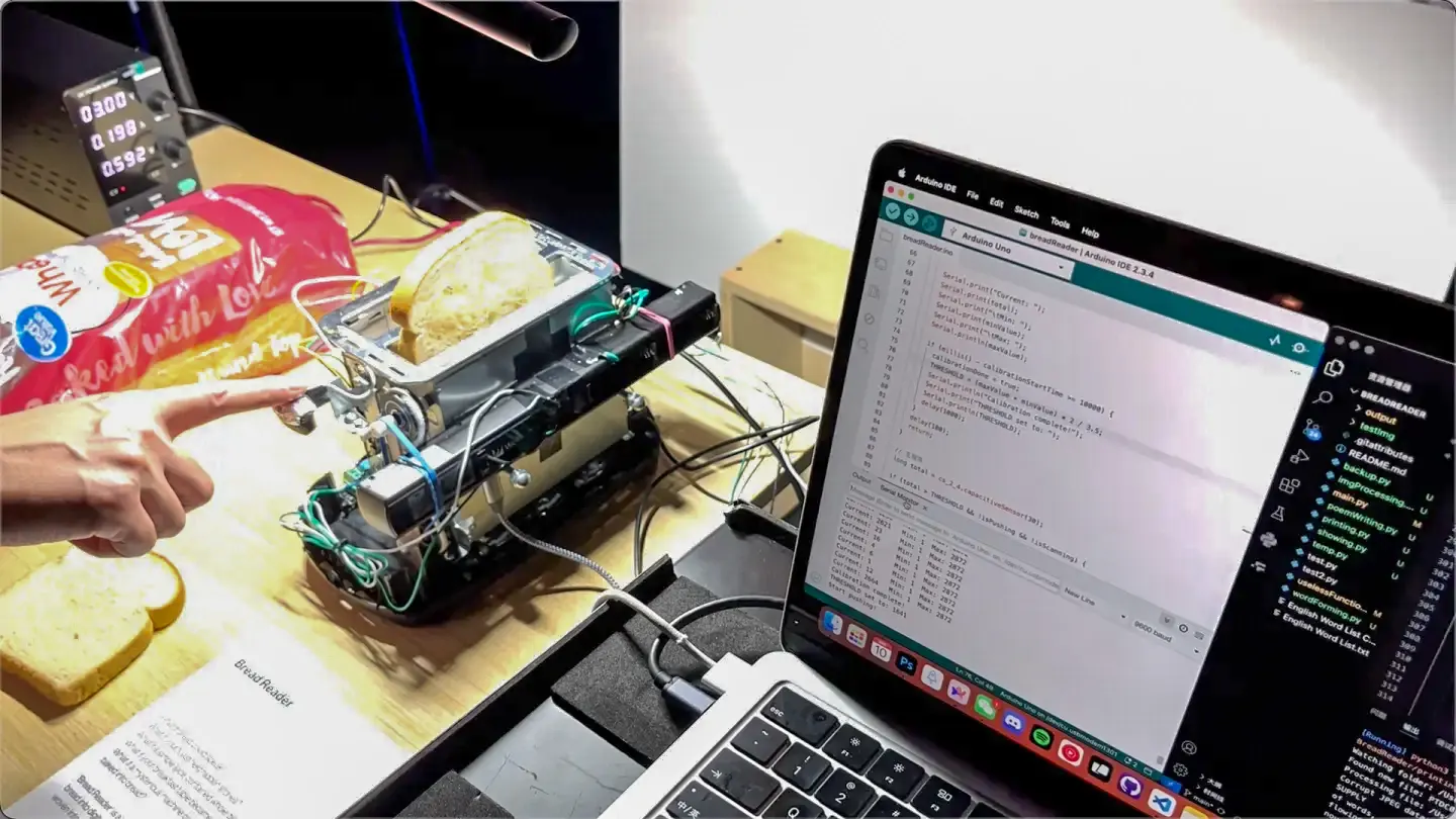 A hand pushes a slice of bread into the machine, a laptop running beside it
